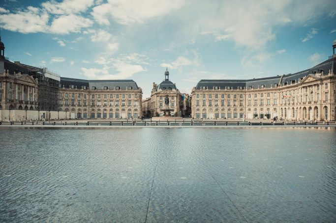 Historic buildings reflect in water under a blue sky with scattered clouds.